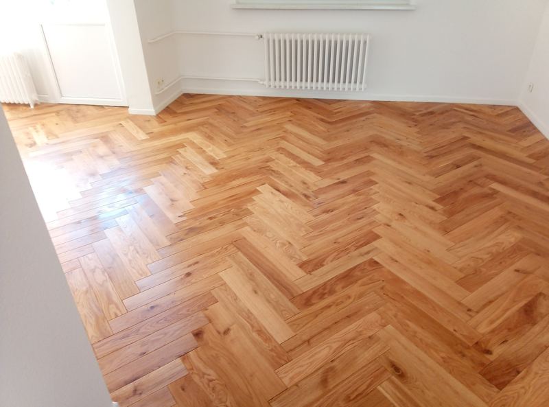 Parquet chêne verni posé en chevrons dans une pièce vide aux murs blancs, radiateur à colonnes