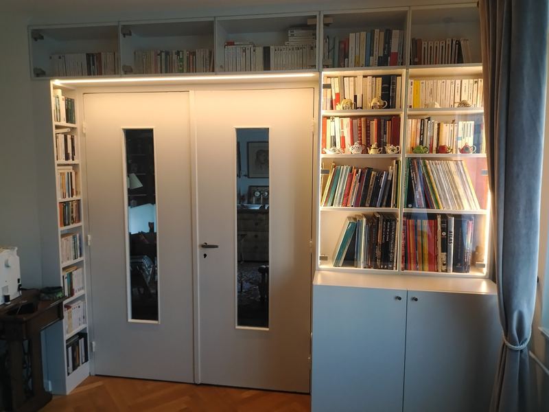 Bibliothèque blanche du sol au plafond encadrant une double porte à vitrages étroits, éclairage LED intégré, meubles bas gris-bleu