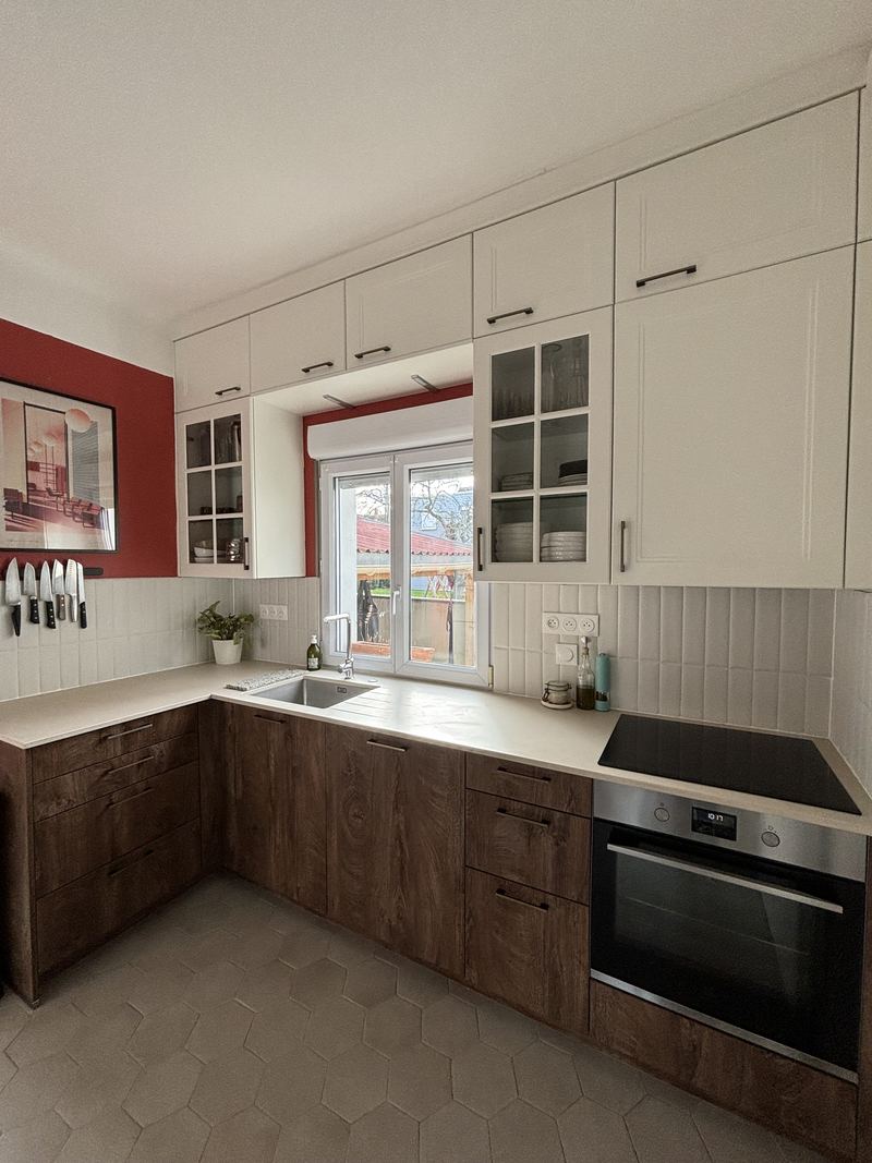 Cuisine en L : meubles bas décor bois foncé veiné à poignées barres noires, meubles hauts crème dont deux vitrés, plan de travail clair, crédence en carreaux blancs verticaux, évier inox sous la fenêtre, plaque induction et four inox