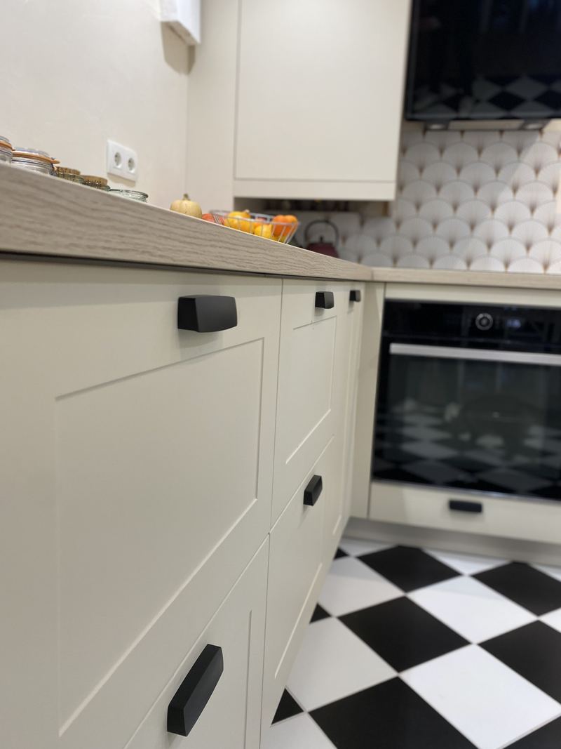Gros plan à arrière-plan flou : façades crème à cadre et poignées noires, plan de travail décor bois clair, crédence en carreaux éventail blancs, four noir