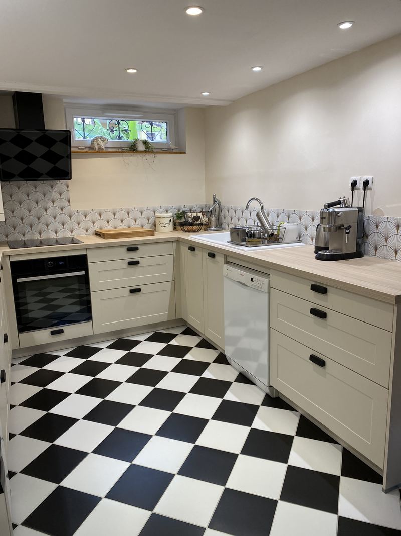 Vue d'ensemble de la cuisine en U crème à cadre (poignées noires), plan de travail décor bois clair, crédence en carreaux éventail, hotte inclinée noire, four noir, lave-vaisselle blanc, spots au plafond