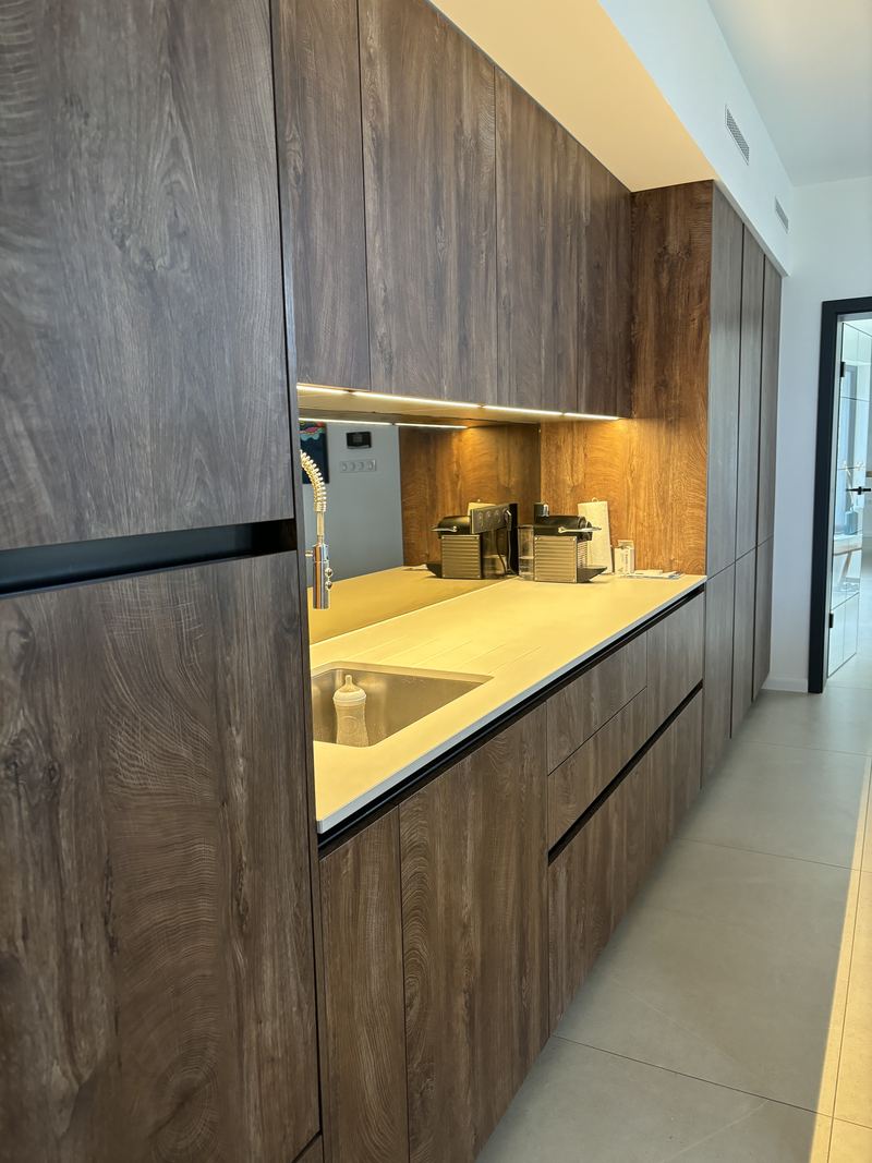 Linéaire sans poignées (gorges noires) en décor bois foncé veiné du sol au plafond, plan de travail blanc, crédence miroir éclairée par LED, évier sous plan et robinet douchette inox