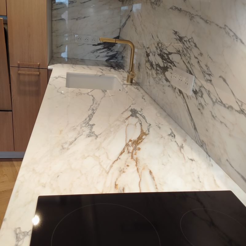 Détail : plan de travail et crédence effet marbre blanc veiné de gris et d'or, évier blanc sous plan, robinet doré, plaque induction noire
