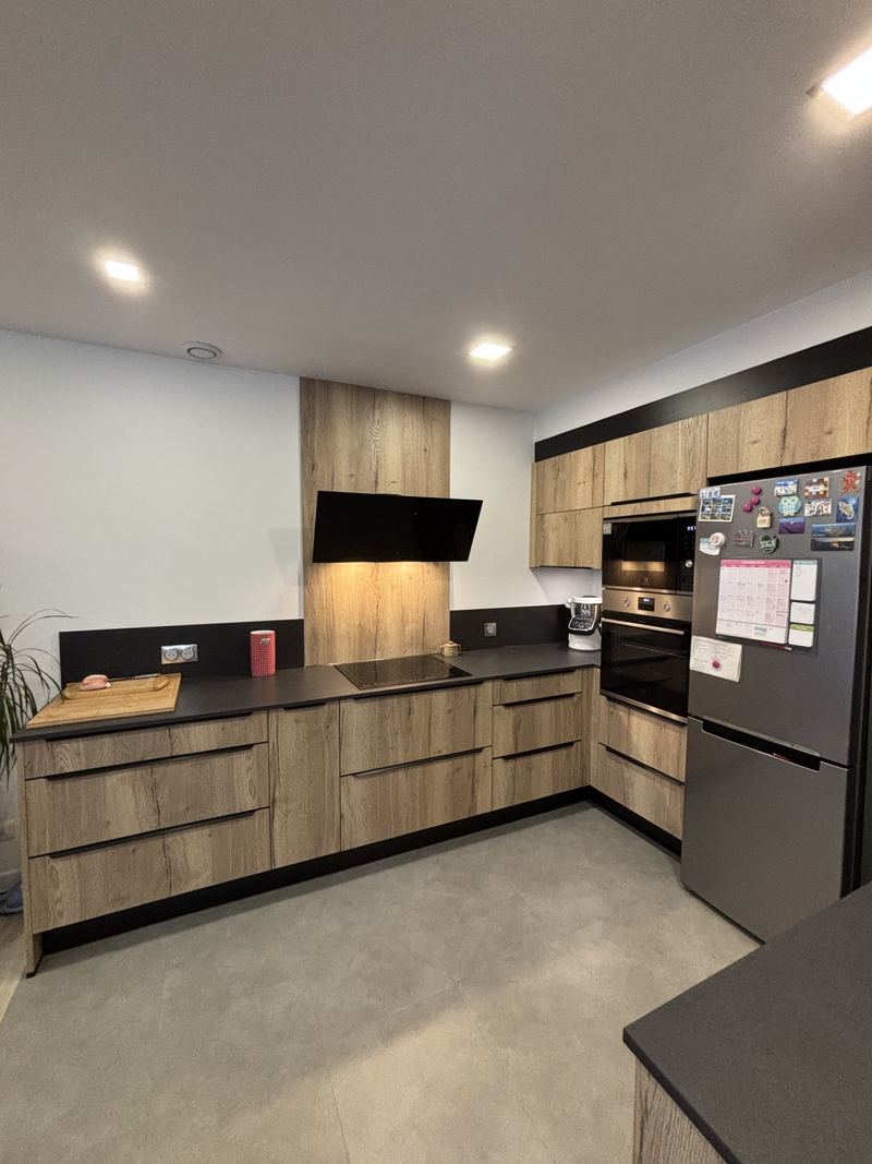 Autre vue de la même cuisine : linéaire décor chêne noueux, hotte inclinée noire sur panneau chêne, plan noir, colonne de fours, réfrigérateur inox