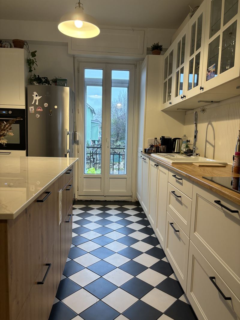 Cuisine en couloir : à droite meubles blancs à cadre et meubles hauts vitrés, plan de travail bois, crédence en lambris blanc