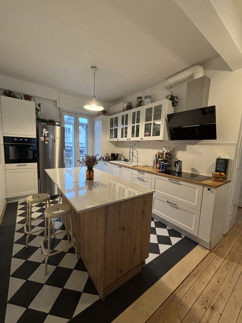 Vue d'ensemble : cuisine blanche à cadre avec meubles hauts vitrés et hotte inclinée noire, îlot central chêne à plan blanc, sol en damier noir et blanc bordé de parquet ancien