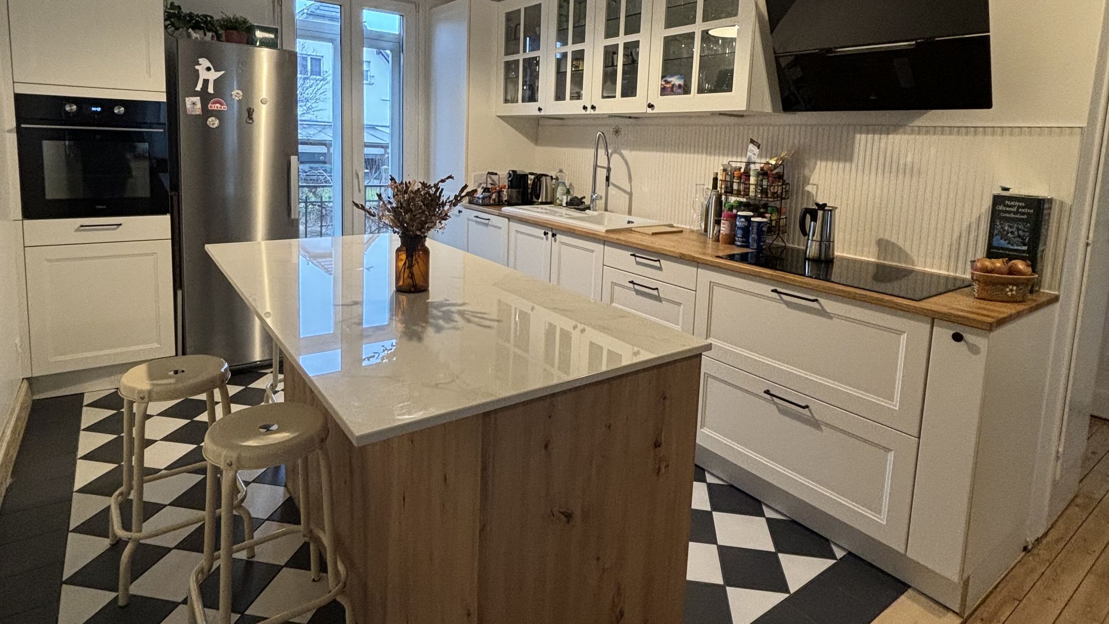 Vue d'ensemble : cuisine blanche à cadre avec meubles hauts vitrés et hotte inclinée noire, îlot central chêne à plan blanc, sol en damier noir et blanc bordé de parquet ancien