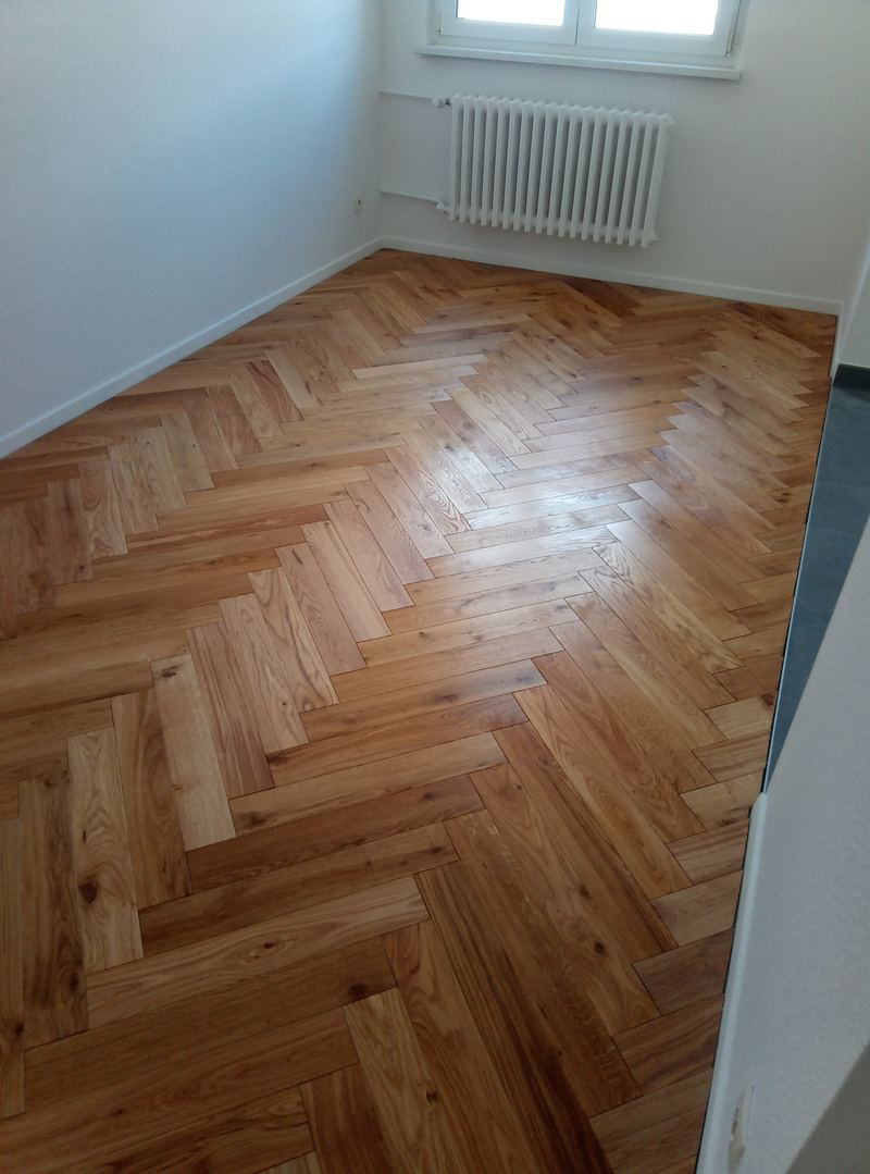 Parquet chêne posé en chevrons dans une pièce étroite aux murs blancs, radiateur à colonnes sous la fenêtre
