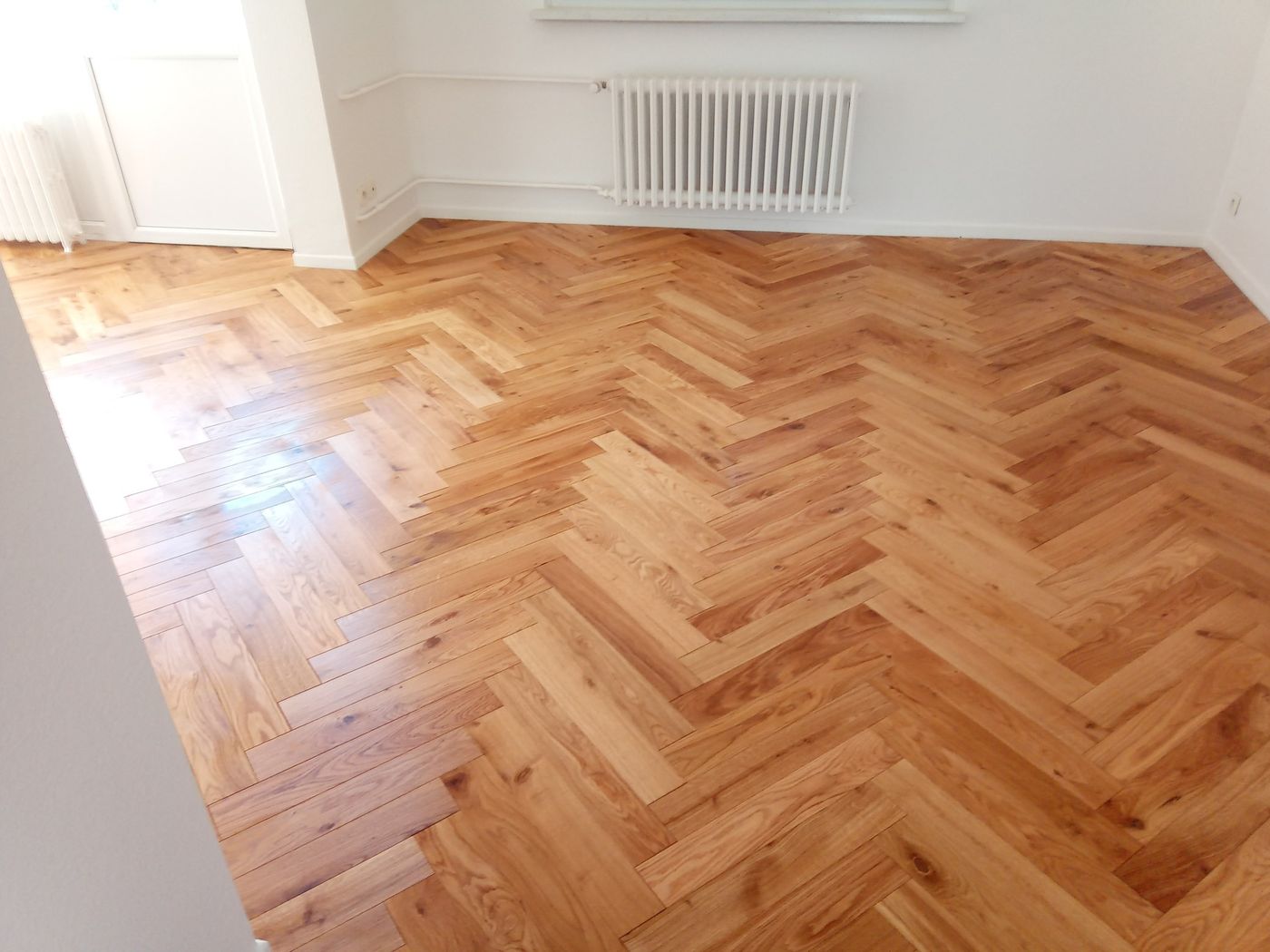 Parquet chêne verni posé en chevrons dans une pièce vide aux murs blancs, radiateur à colonnes
