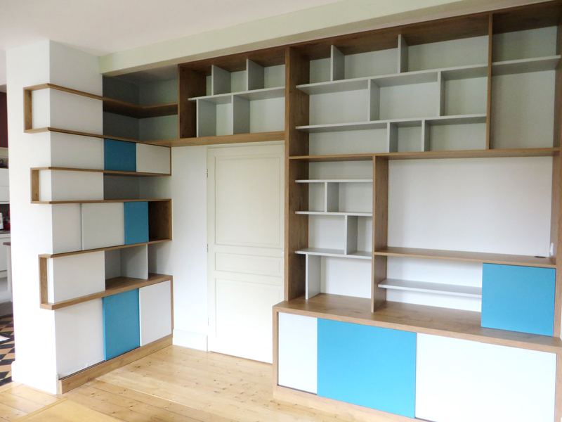 Bibliothèque murale sur mesure encadrant une porte : casiers blancs à chants chêne, étagères décalées, portes coulissantes bleu turquoise et blanches en partie basse