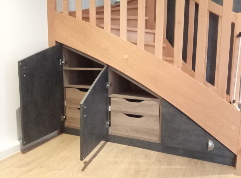 Rangement sous escalier : portes décor ardoise sombre ouvertes sur tiroirs et étagères décor chêne, sous un escalier bois à balustres