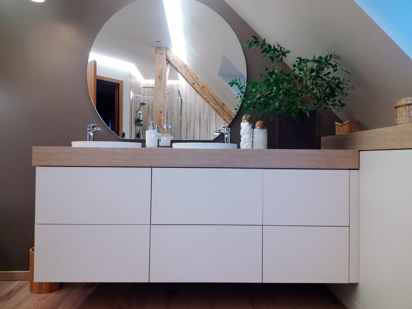 Meuble double vasque suspendu blanc à tiroirs sans poignées, plan chêne, deux vasques posées, grand miroir rond sans cadre, poutres apparentes sous rampant, plante verte