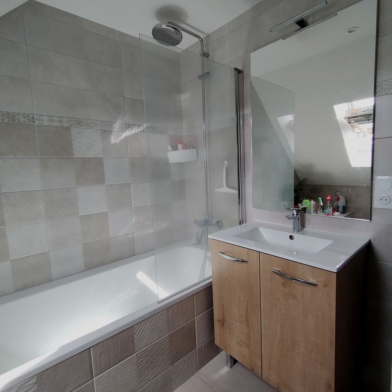 Salle de bains sous toit : baignoire avec pare-bain vitré et colonne de douche, carrelage gris aspect pierre, meuble vasque décor chêne à deux portes, grand miroir, fenêtre de toit